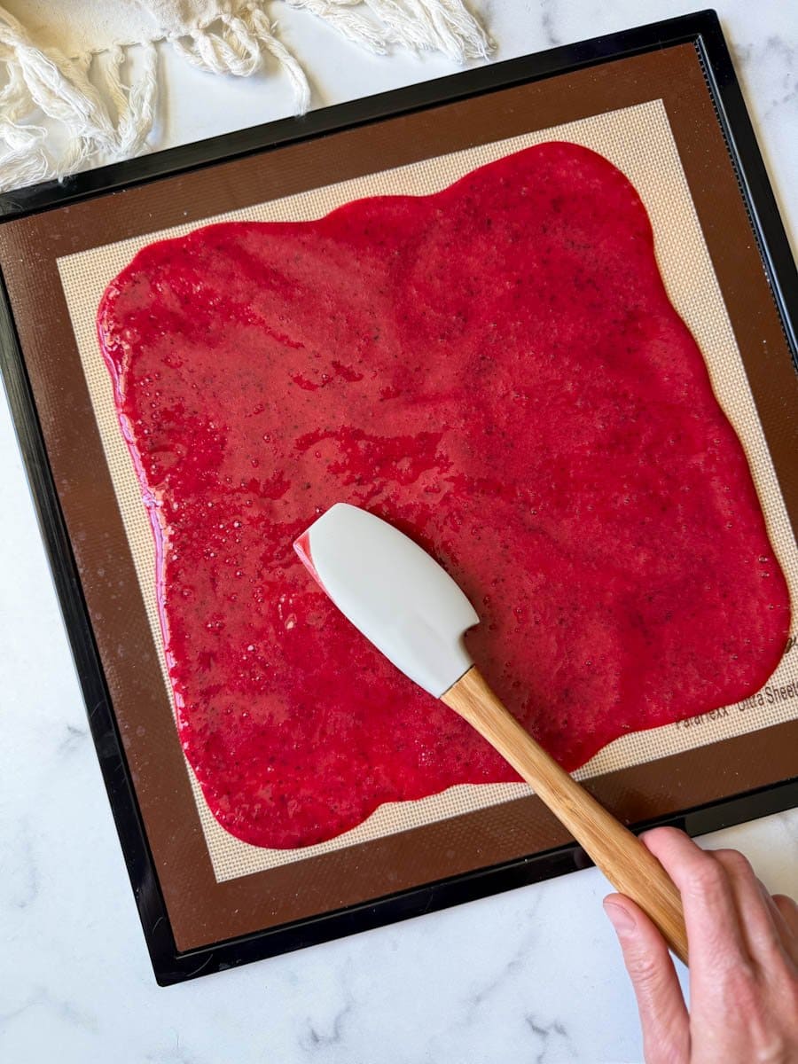 Fresh fruit puree is being spread evenly across a silicone mat with a  spatula. 