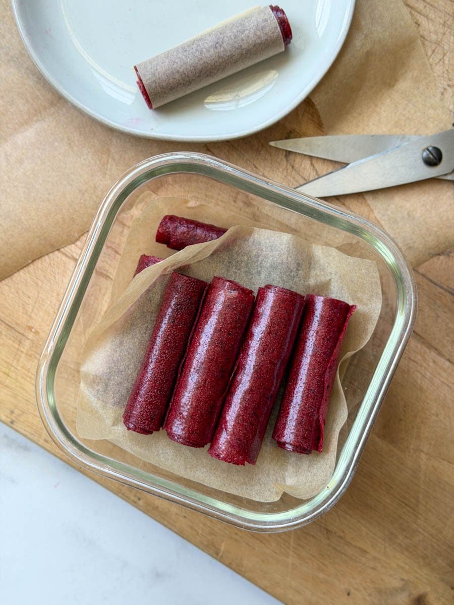 A square glass storage container has fruit leather rolled into individual rolls. A piece of parchment paper is below the top layer with more fruit leather below. 