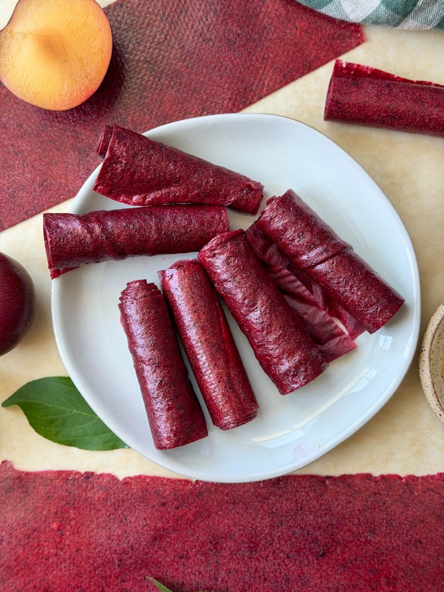 Dried fruit leather rolled into single rolls sit on top of a small white ceramic plate. A few sections of fruit leather and fresh plums are arranged around the plate. 