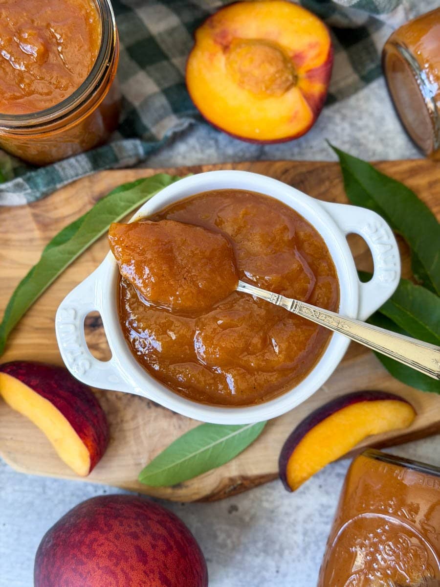 A small white ramekin is full of peach butter, a full spoonful of a silver spoon rests on top of the butter. Slices of fresh peaches and leaves surround the ramekin along with a few jars of peach butter from canning. 