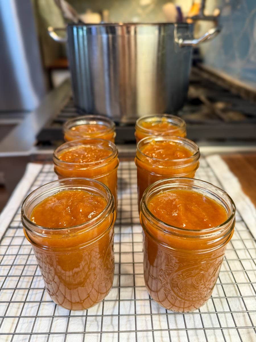 Six half pint jars full of peach butter are filled to 1/4 inch head space sit on top of a wire cooling rack, ready for canning. 