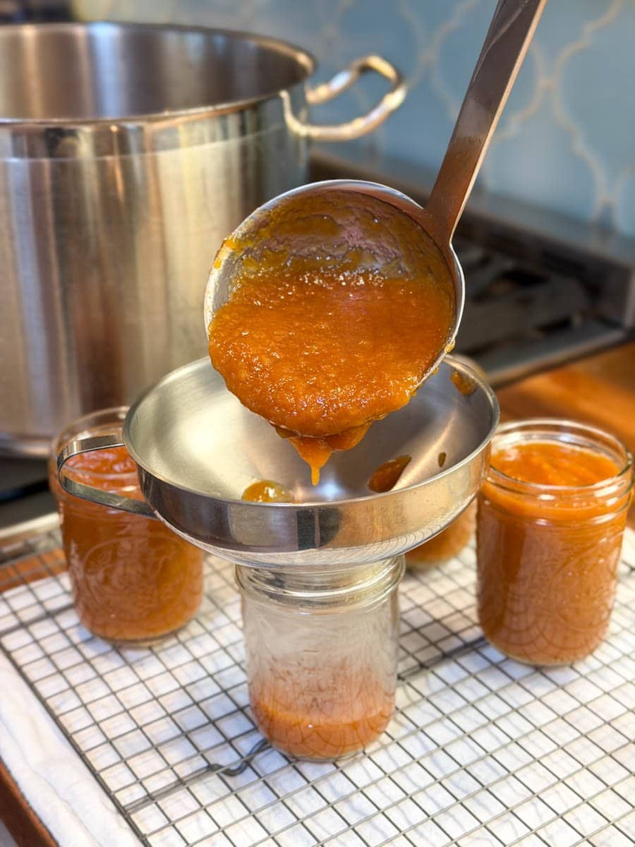 A stainless steel ladle is being used to fill half pint jars of fruit preserves with a canning funnel. A large stock pot is on the stove beyond. 