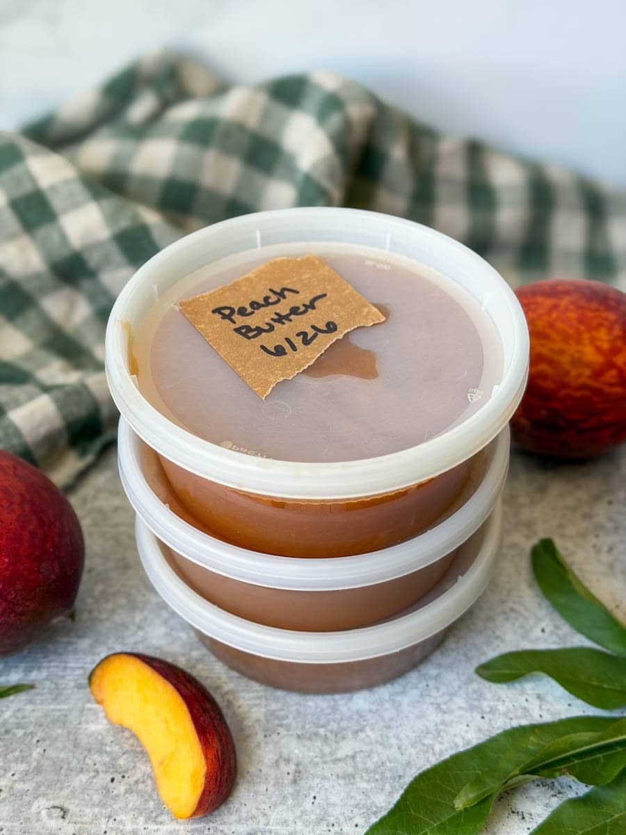Three freezer safe plastic containers are full of peach butter with one of the lids being labeled as such. Two whole peaches are next to the containers. 