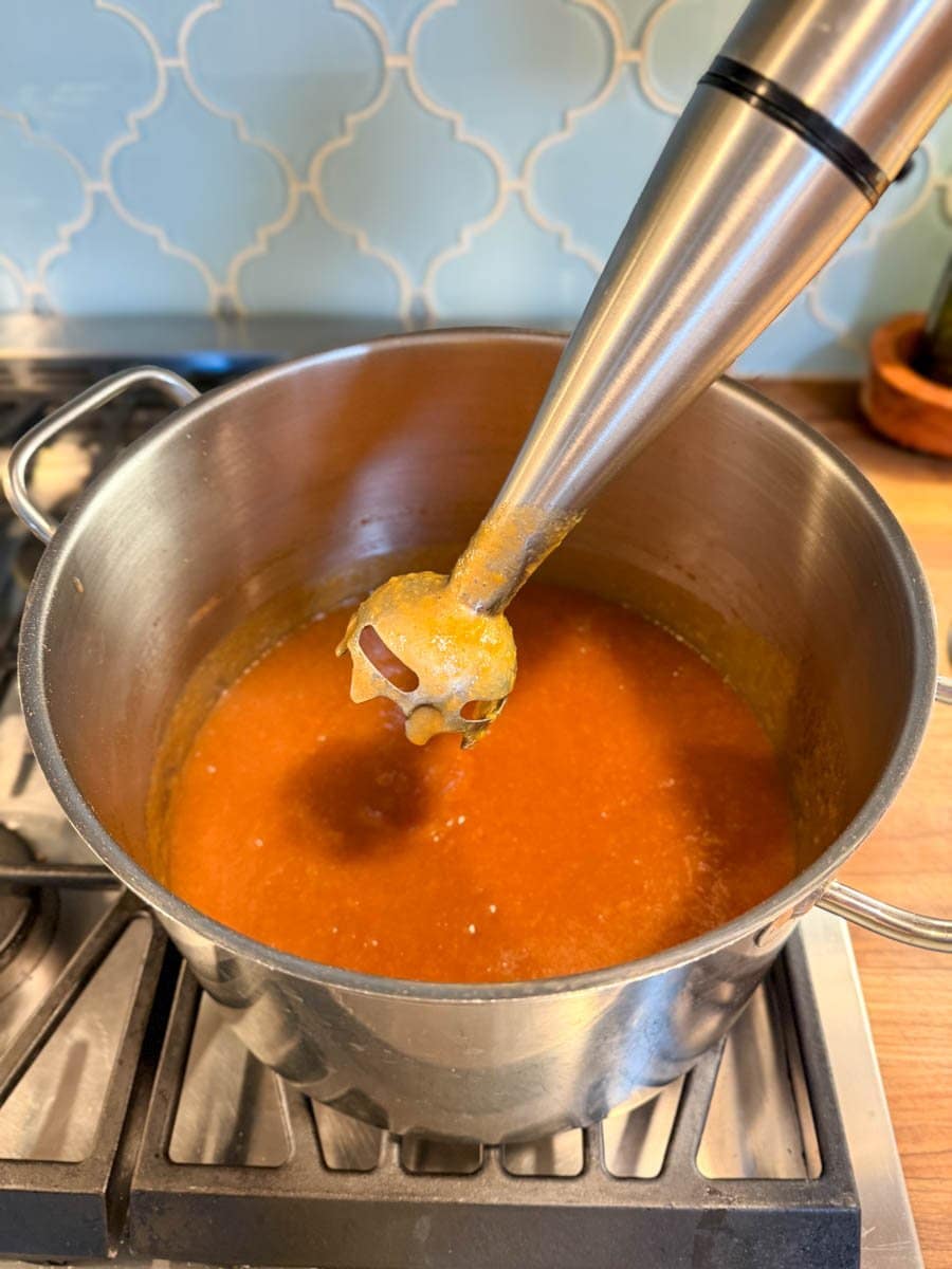 An immersion blender is held above a stock pot full of peach butter after blending. 