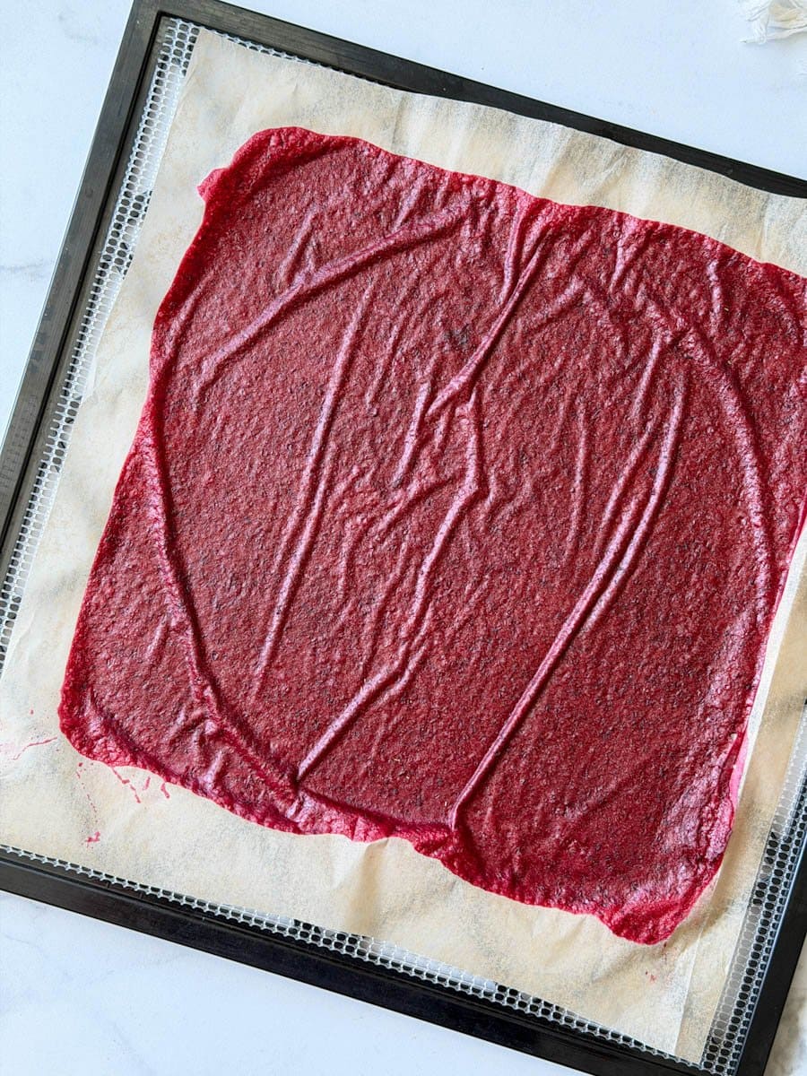 Dried fruit leather that is purple to red in color is on top of a dehydrator tray lined with parchment paper. 