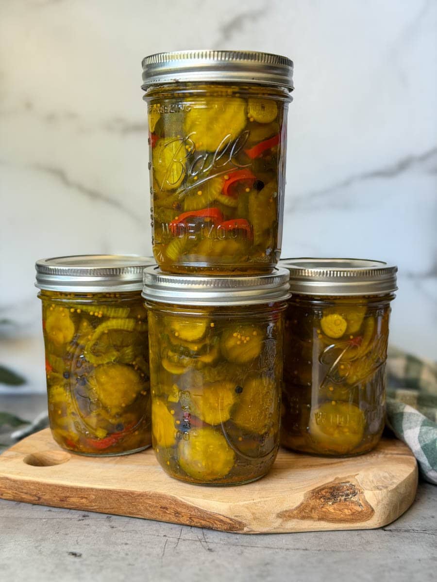 Three pint jars of bread and butter pickles are lined up on a small wood cutting board with another jar stacked on top of the one in the middle.
