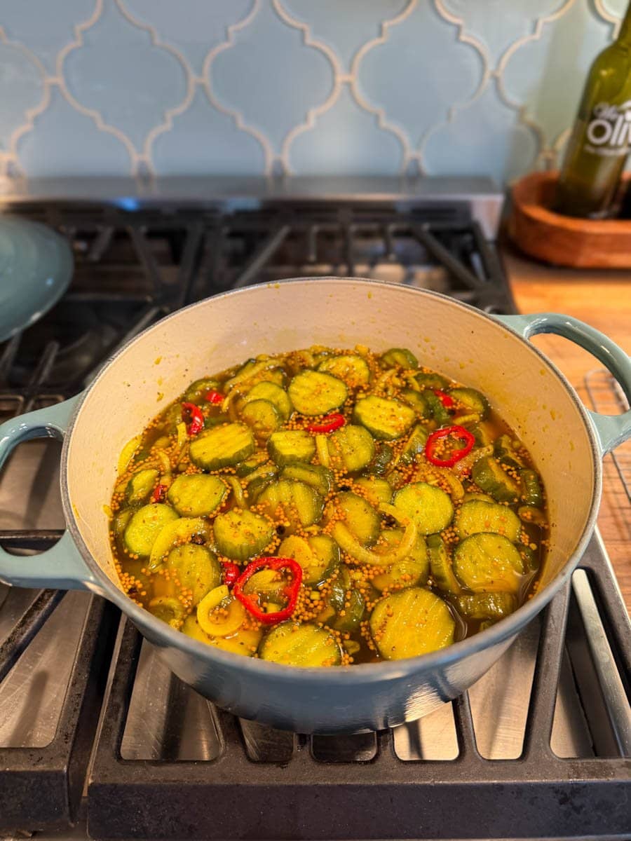 A pot on the stove of bread and butter pickle mixture with the brine. Cucumber, sliced onions, peppers, and mustard seeds are most visible.