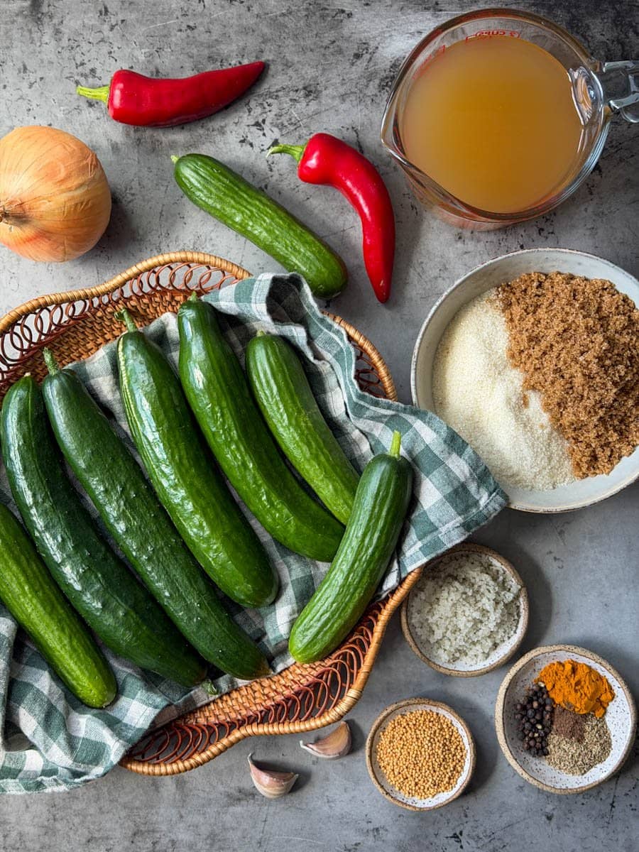 A wicker basket of fresh cucumbers with a glass liquid measuring cup of vinegar, a bowl of white and brown sugar, along with various spices, salt, fresh garlic, peppers, and onions arranged around it.
