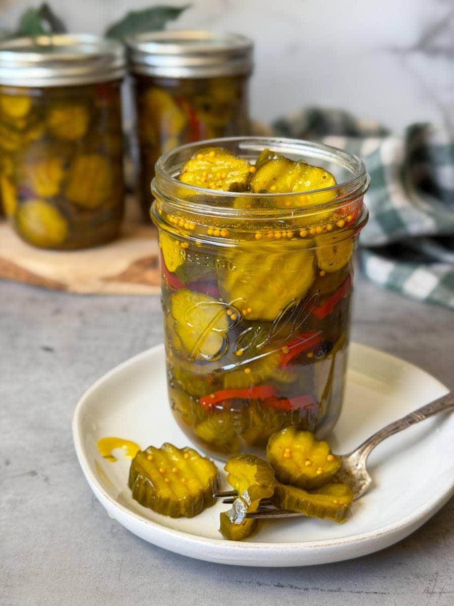 Bread and butter pickles in a pint jar sitting on a small ceramic plate. A few of the pickles are on the plate with a fork nearby. Beyond are more jars of finished bread and butter pickles.