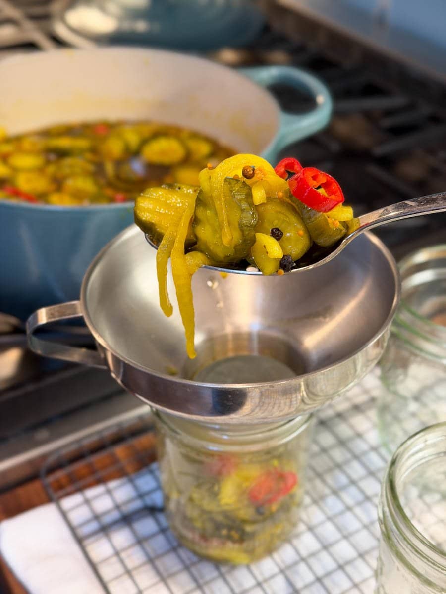 A slotted spoon full of the cucumber mixture is held above a jar which is halfway full with a canning funnel on top of it. Beyond is a pot on the stove with the rest of the cucumber mixture and brine.
