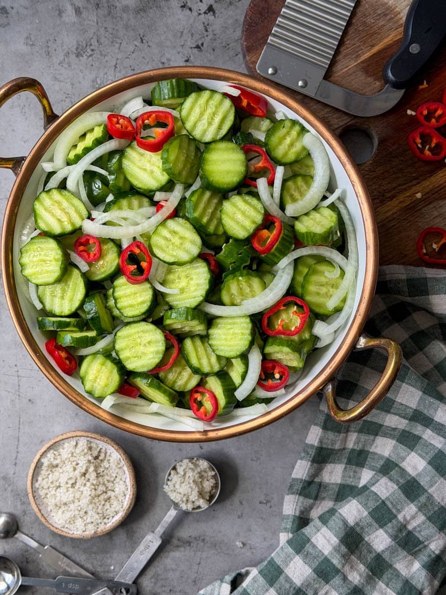 A white ceramic bowl full of a mixture of cucumber slices, onions slices, and peppers slices. A wood cutting board is nearby with a crinkle cutter and sliced peppers on top of it.