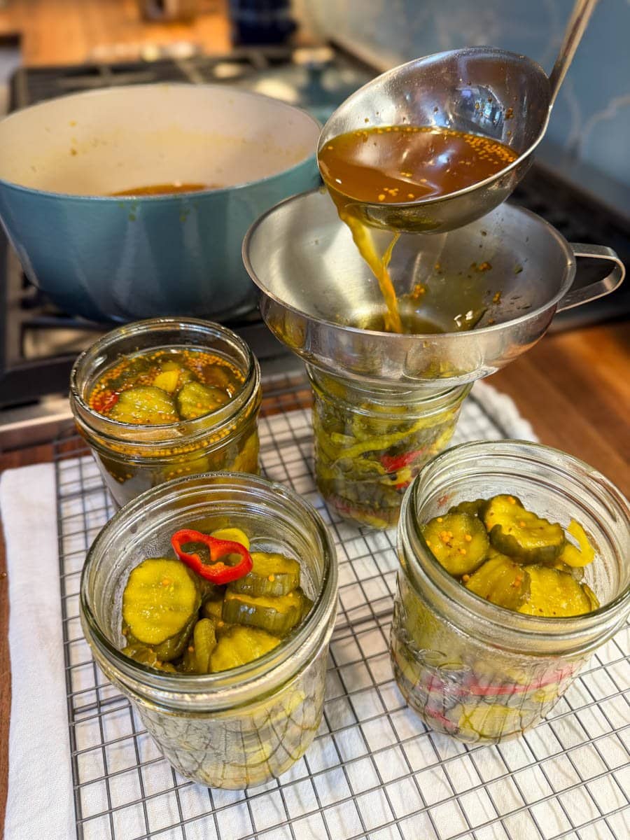 A metal ladle is pouring brine into the jars already filled with the cucumber mixture using a canning funnel.
