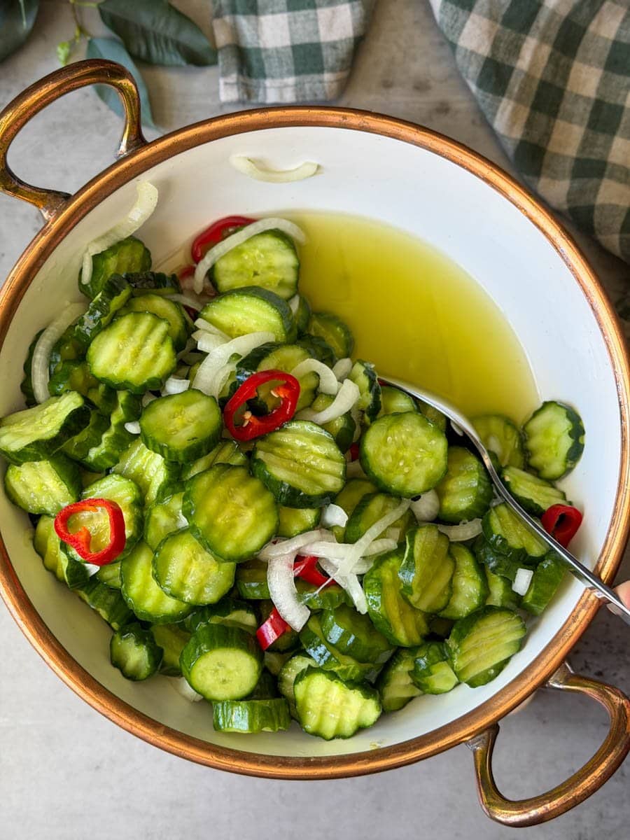 A white ceramic bowl is full of the salted cucumber mixture with onions and peppers. A metal spoon is being used to pull aside the mixture to reveal green watery liquid beneath.