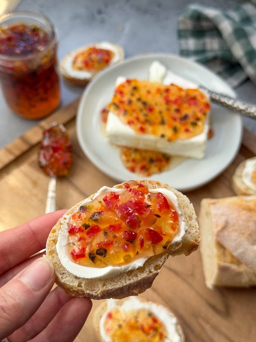A small slice of bread with a cream cheese smear has a dollop of pepper jelly on top of it. Below on a wood cutting board is the rest of the loaf of bread along with cream cheese topped with pepper jelly.