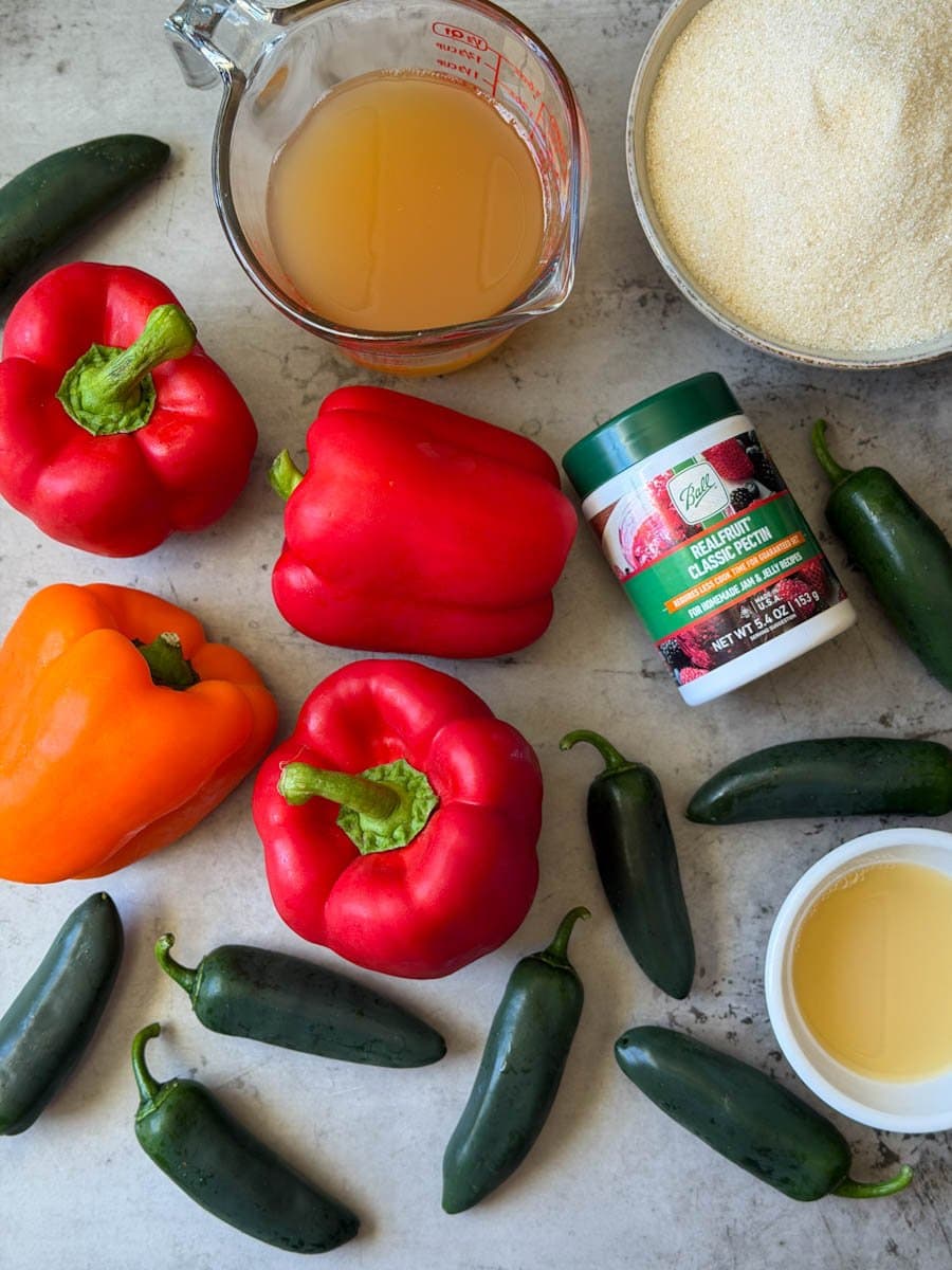 A variety of chilis, pectin, sugar, lime juice and apple cider vinegar are arranged on a counter.