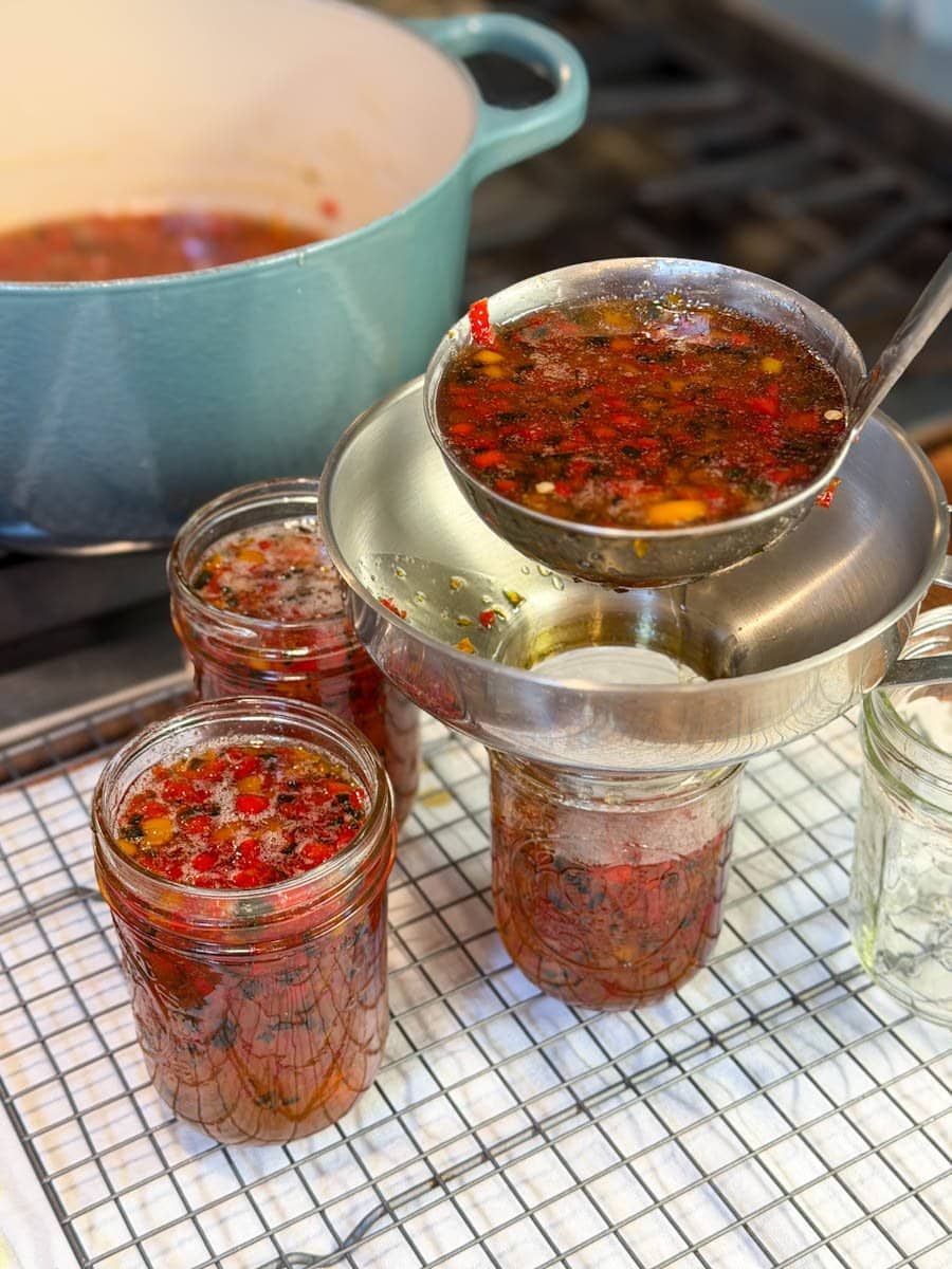 A metal ladle is spooning hot pepper jelly into half pint mason jars on a wire cooling rack via a canning funnel.