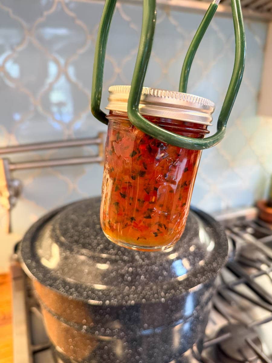 A jar lifter is holding a full half pint mason jar in front of a large canning pot on the stove.