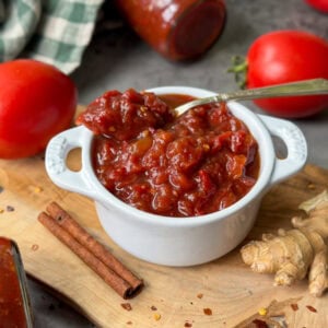 A white ramekin full of spiced tomato jam. Spoon is placed inside the jam and there are a few whole tomatoes, cinnamon sticks, ginger, and red chili flakes surrounding the jar.