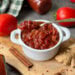 A white ramekin full of spiced tomato jam. Spoon is placed inside the jam and there are a few whole tomatoes, cinnamon sticks, ginger, and red chili flakes surrounding the jar.