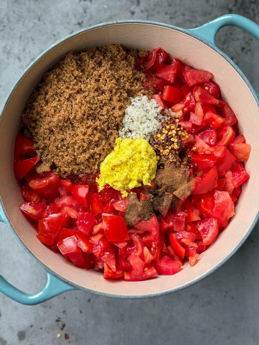 A large pot is full of chopped tomatoes, chopped ginger, brown sugar, salt, cinnamon, red chili flakes, and all spice. 