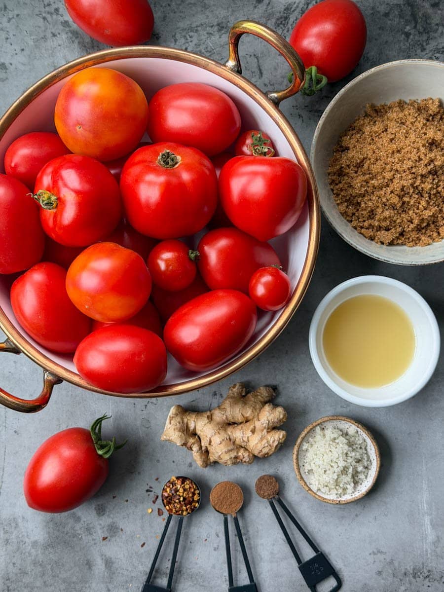 A bowl full of fresh tomatoes is surrounded by a bowl of brown sugar, and small ramekin of lime juice, fresh ginger, salt, and various spices. 