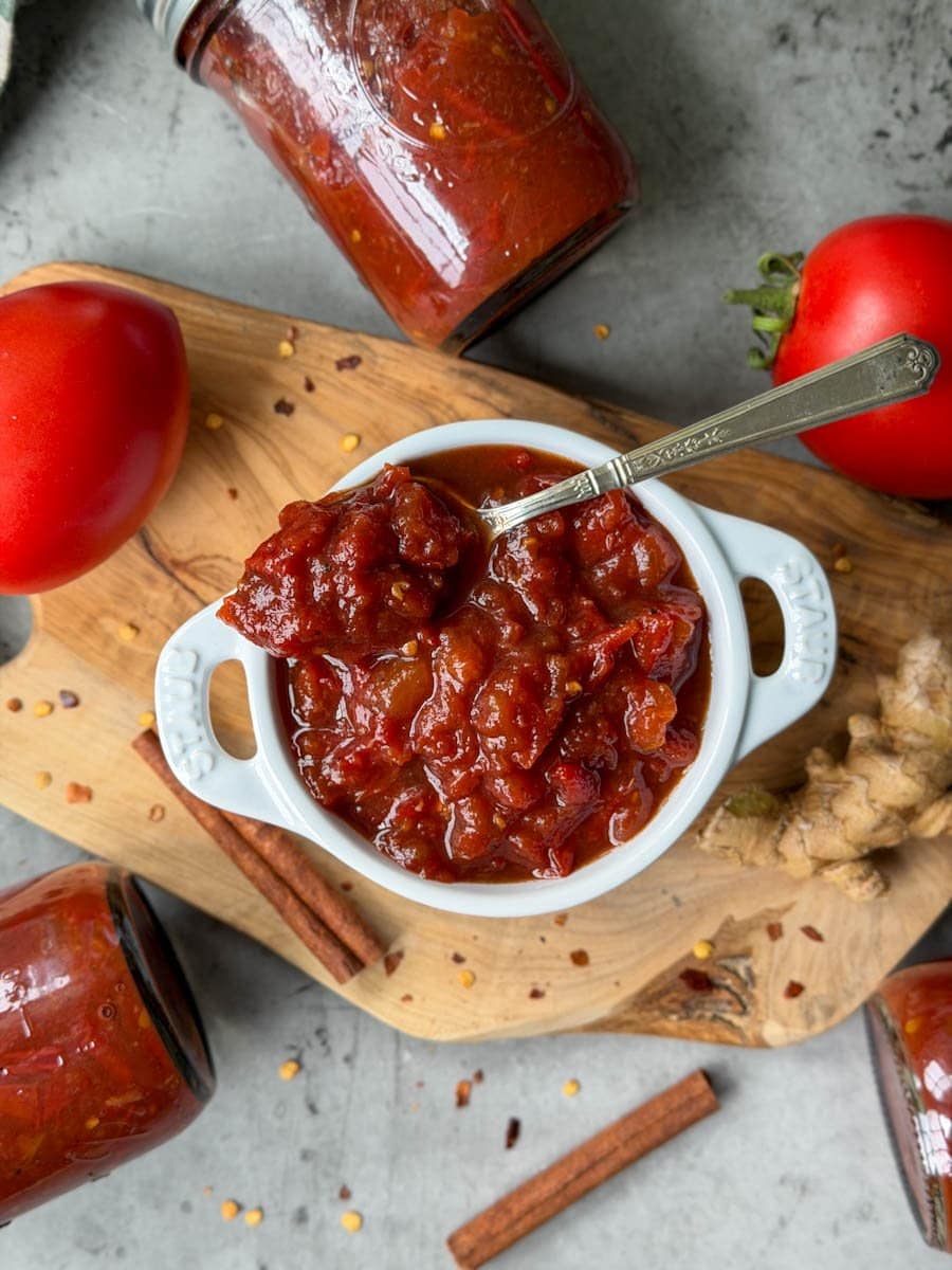 A white ramekin is full of tomato jam, a spoon is inserted into the jam. Surrounding the ramekin are fresh tomatoes, cinnamon sticks, fresh ginger, red chili flakes, and a few jars of tomato jam. 