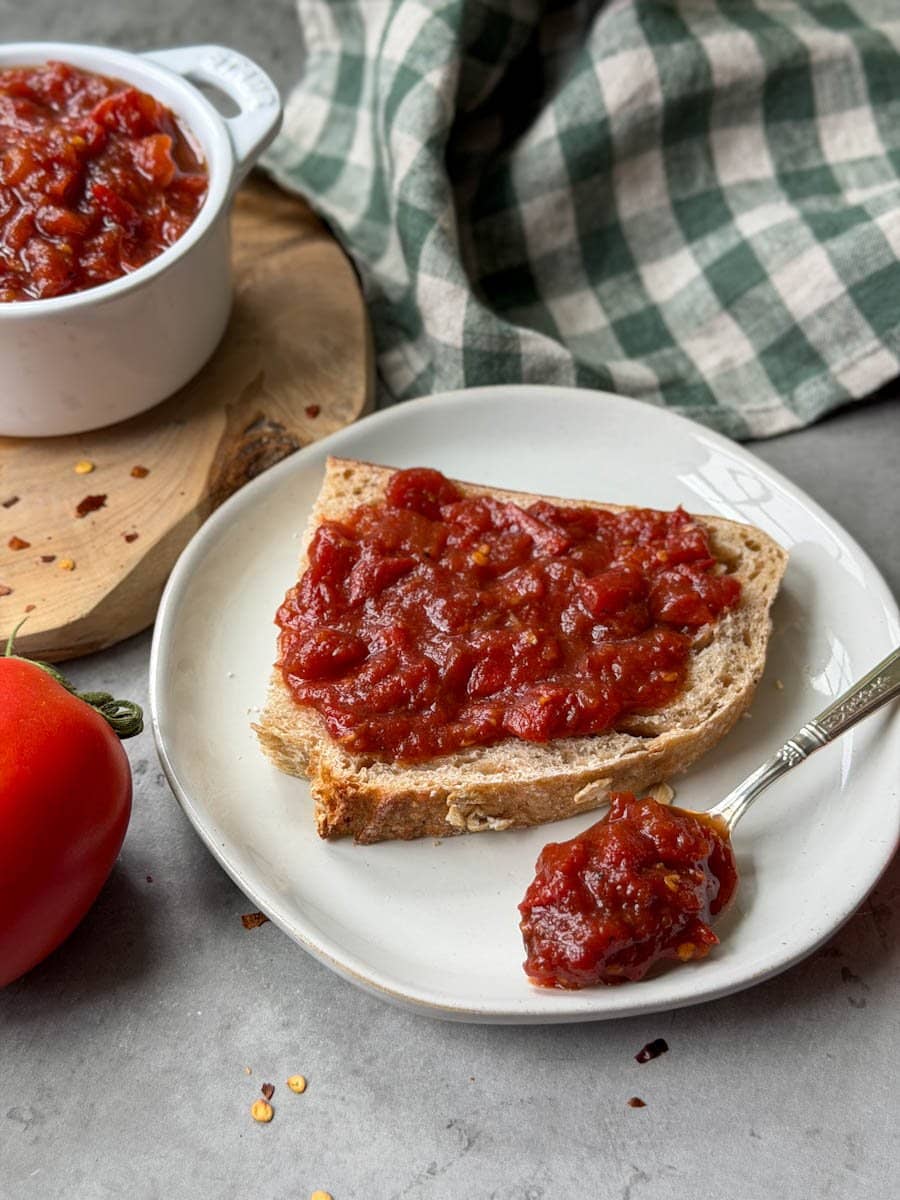 A slice of bread on a small white plate has tomato jam spread across the top of the slice while a spoon that is full of tomato jam rests along the side of the bread.