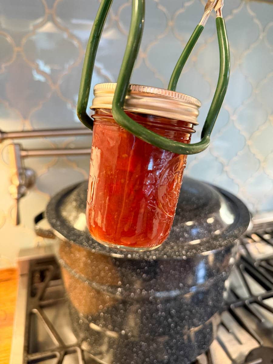 A half pint mason jar is being held with a jar lifter in front of a canning pot. 
