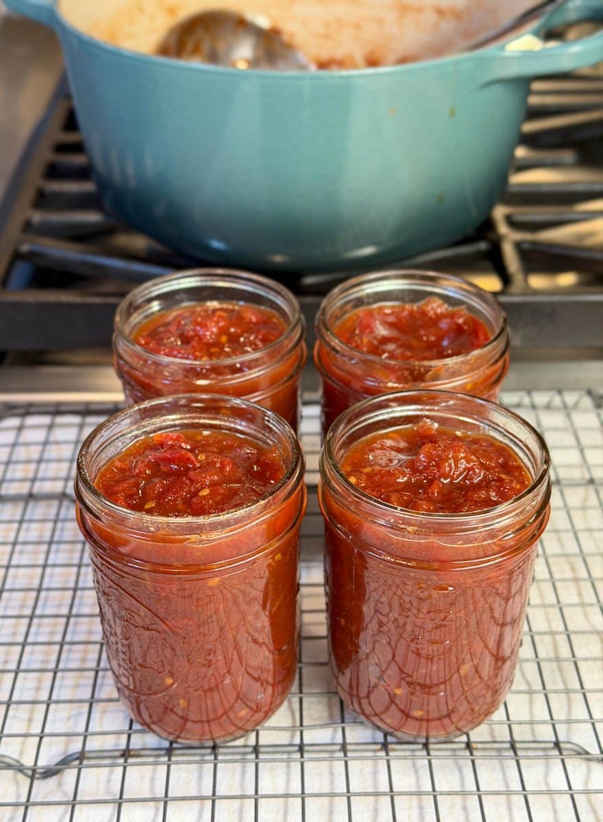 Four half pint jars full of tomato jam sit on top of a wire cooling rack in front of a heavy bottom pot on the stove in which the jam was cooked. 