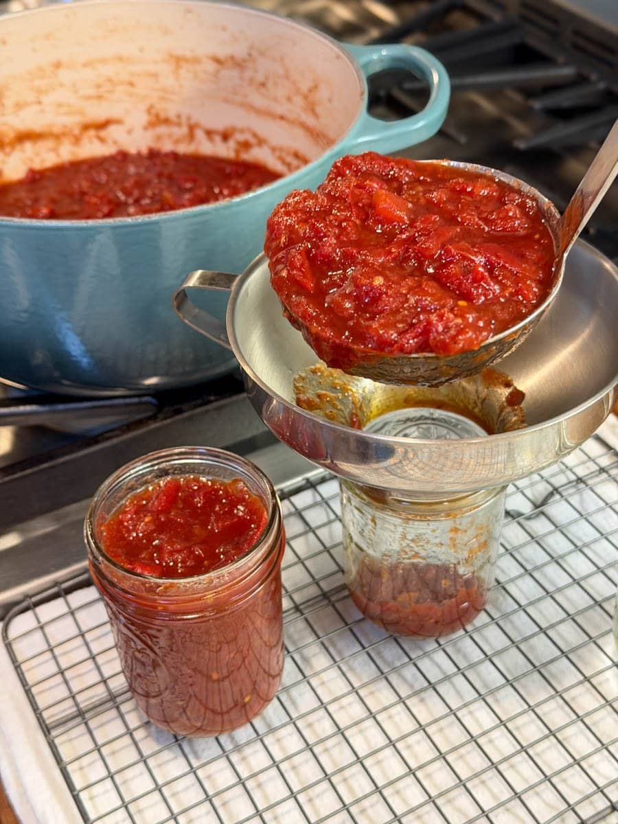 A ladle is being used to fill half pint mason jars via a canning funnel. A large pot is on the stove full of the preserves. 