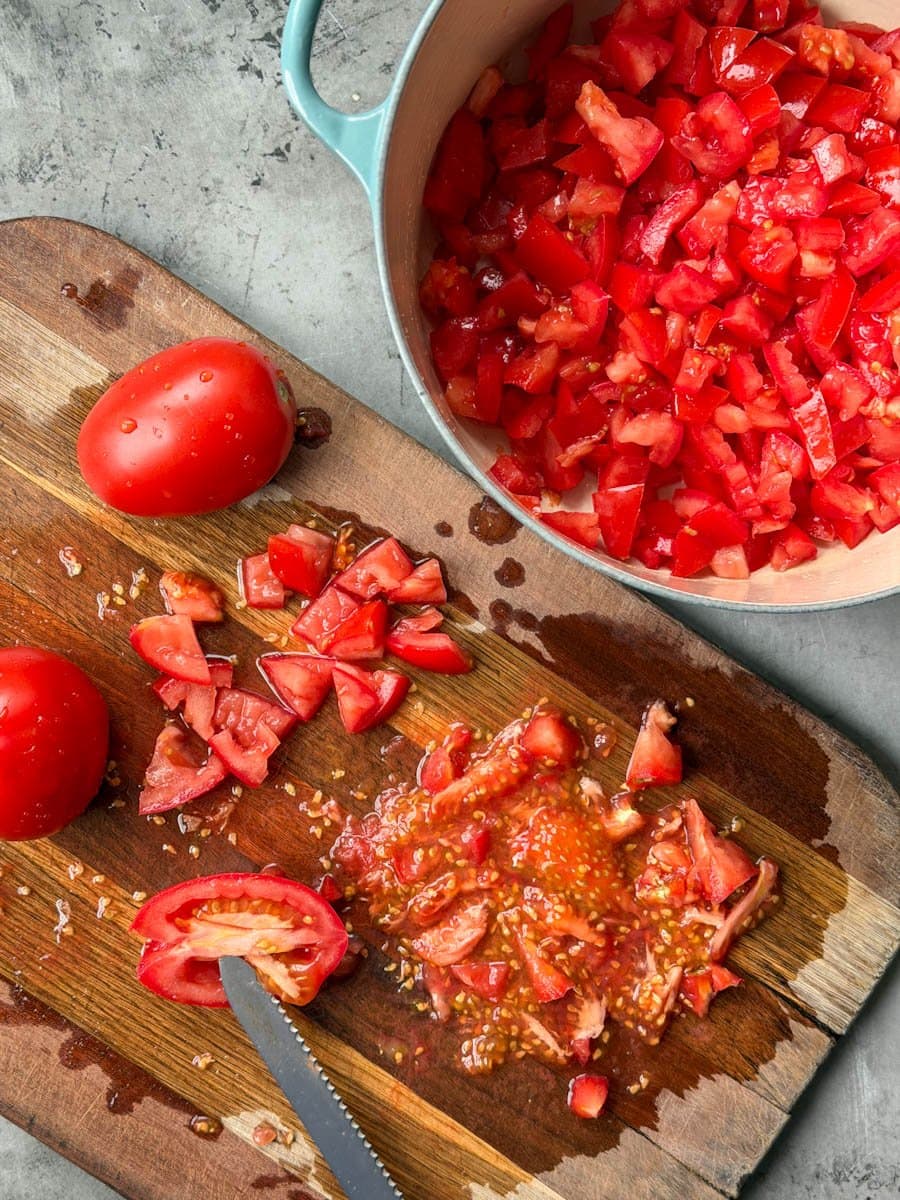 Fresh veggies are being chopped on a wood cutting board. Some of  the seeds are being removed during the process as the chopped ingredients are put into a large pot. 