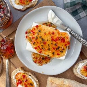 A ceramic plate with a block of cream cheese on top of hit has been smothered in pepper jelly. Slices of bread surround the plate which contain cream cheese and pepper jelly on top.