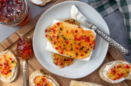 A ceramic plate with a block of cream cheese on top of hit has been smothered in pepper jelly. Slices of bread surround the plate which contain cream cheese and pepper jelly on top.