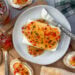 A ceramic plate with a block of cream cheese on top of hit has been smothered in pepper jelly. Slices of bread surround the plate which contain cream cheese and pepper jelly on top.