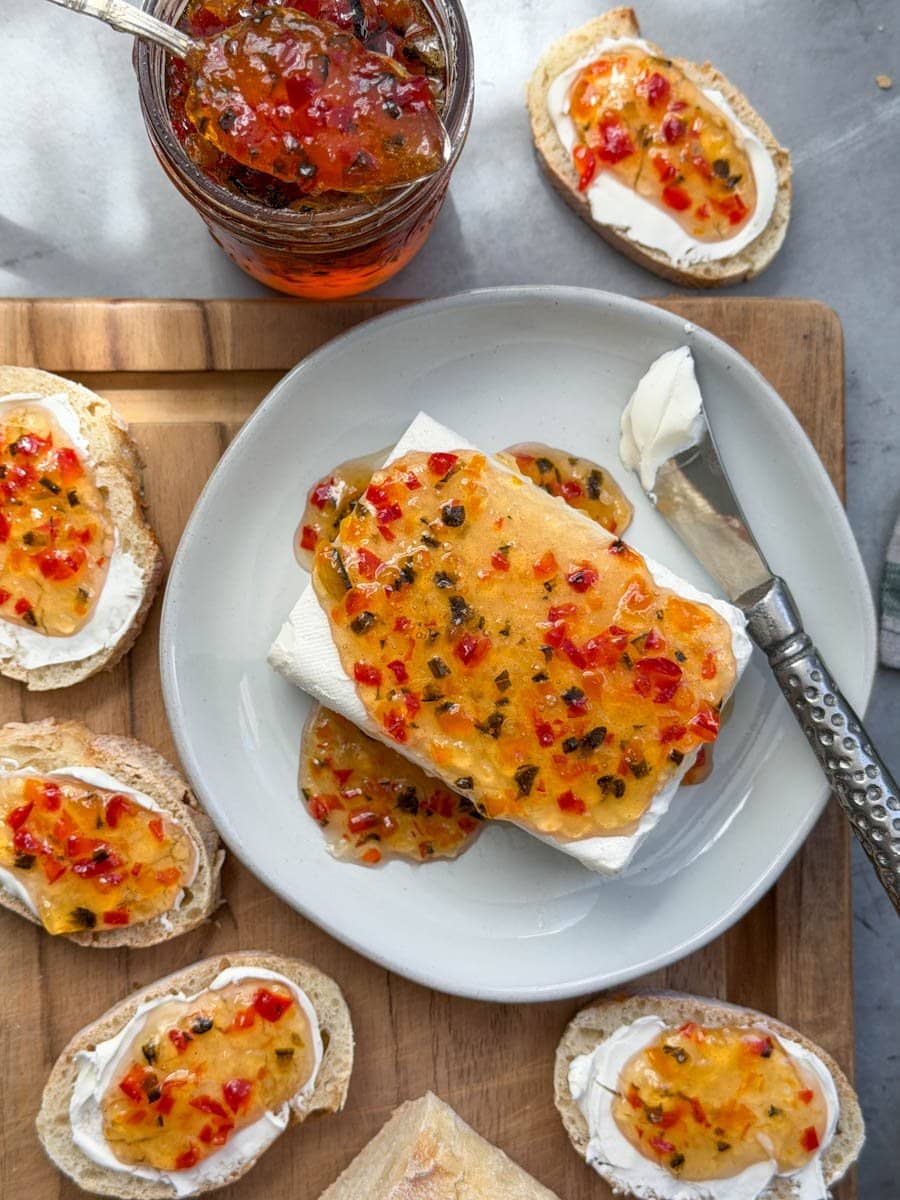 A block of cream cheese sits on a small ceramic plate with pepper jelly poured over the top of it, spilling out over the sides. Surrounding the plate are slices of a baguette with cream cheese and pepper jelly on top of them.