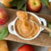 A ramekin full of homemade applesauce. A spoon is resting on the top of the ramekin with a spoonful of the applesauce. A few fresh apples and jars of applesauce surround the ramekin.