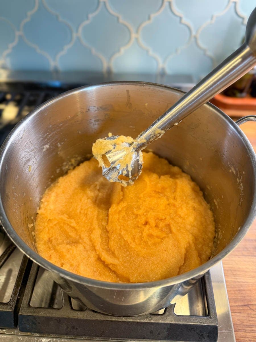 An immersion blender is poised above a stock pot full of blended, cooked, preserves.