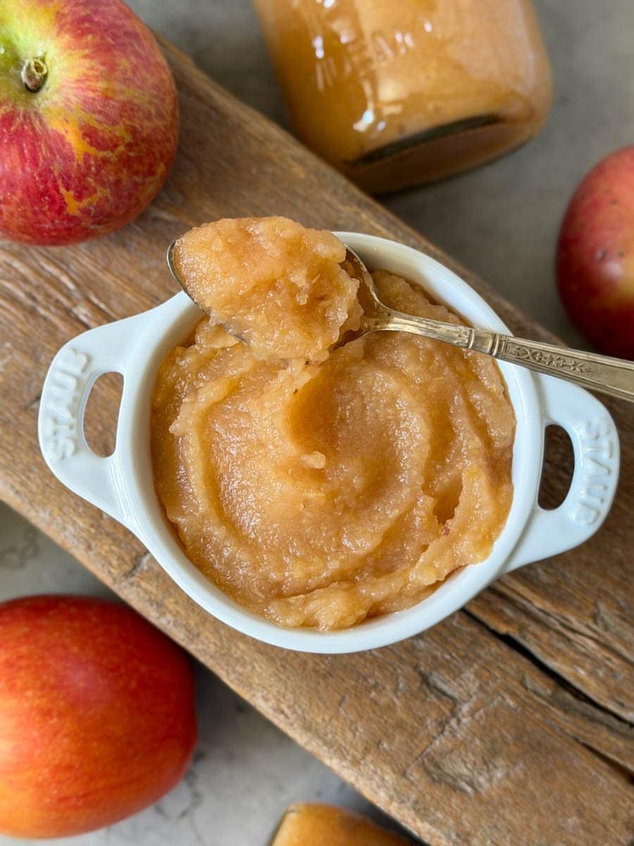 Homemade applesauce fills a small white ramekin while a spoon rests on top full of applesauce. A few fresh apples and jars of applesauce surround the ramekin.