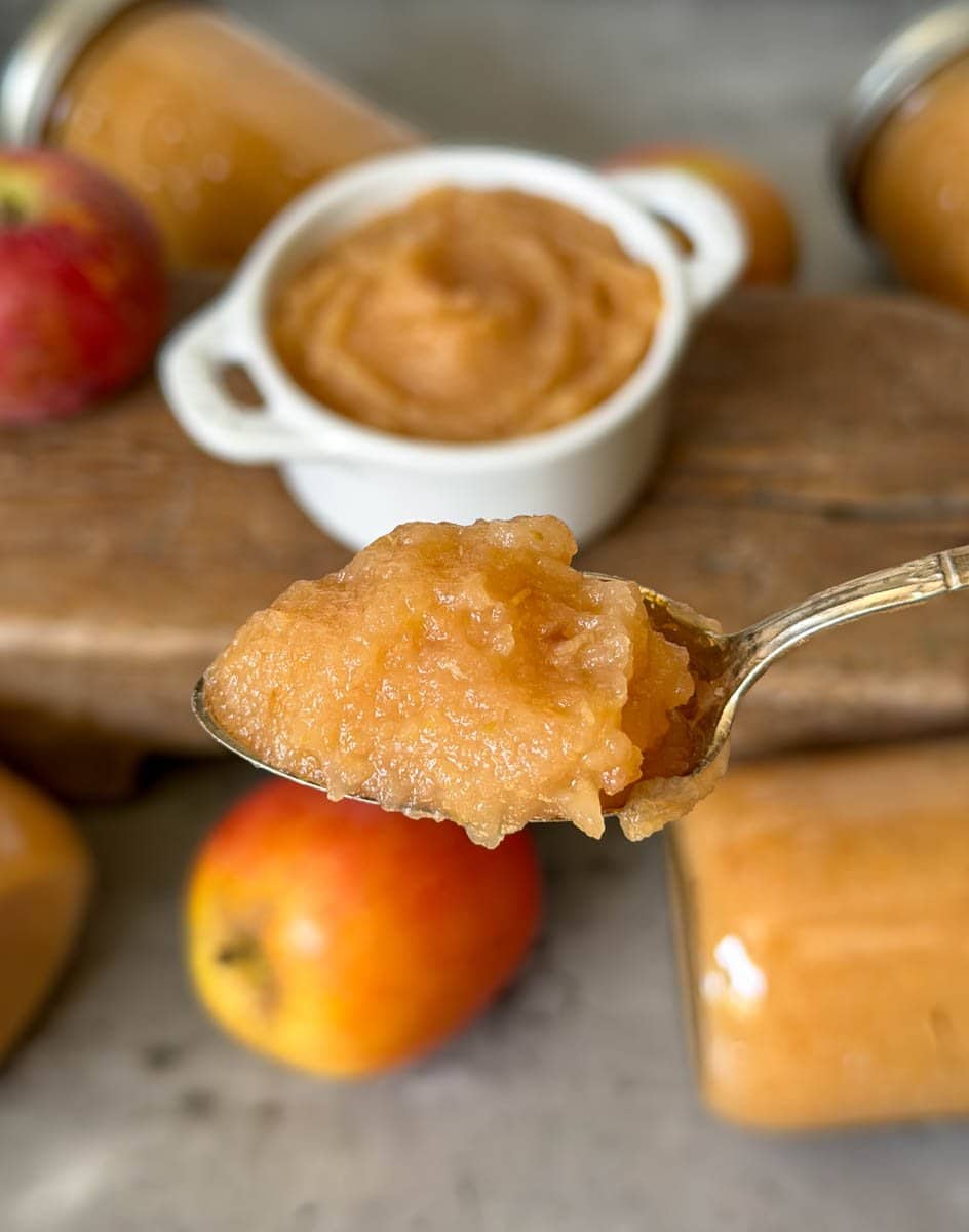 A spoonful of applesauce is held a loft. Beyond is a ramekin full of applesauce, jars of applesauce, and a number of fresh apples.