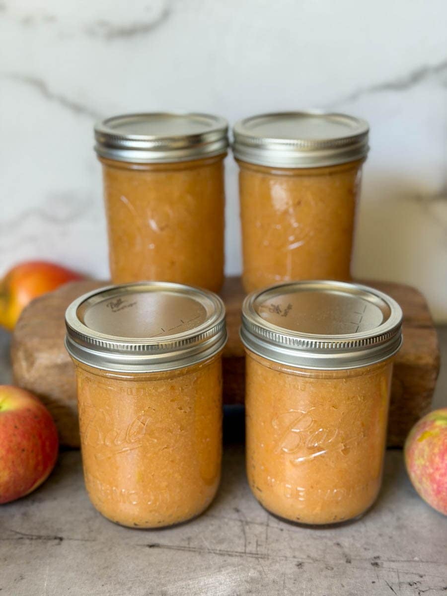 Four pint jars of applesauce are stacked two by two, one row in front of the other. Fresh apples are scattered around the jars.