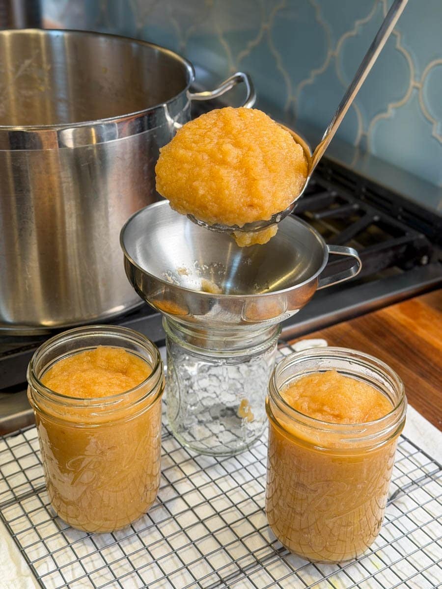 A ladle is being used to spoon applesauce into pint jars using a canning funnel. A large stock pot is in the background on the stove.