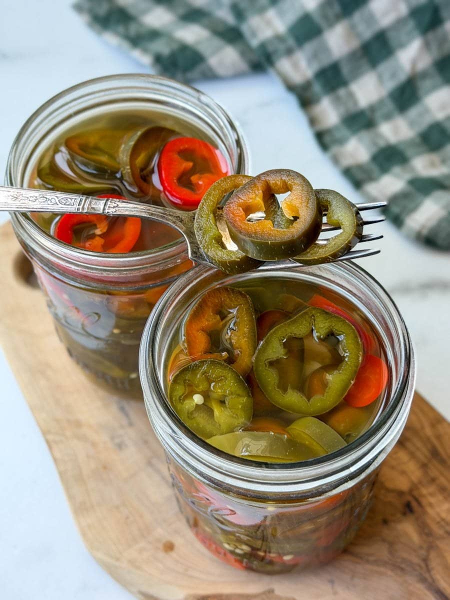 Two pint jars full of sliced pickled jalapeños sitting on a wood cutting board. A fork rests on top of the jars with a few slices on the fork tines.