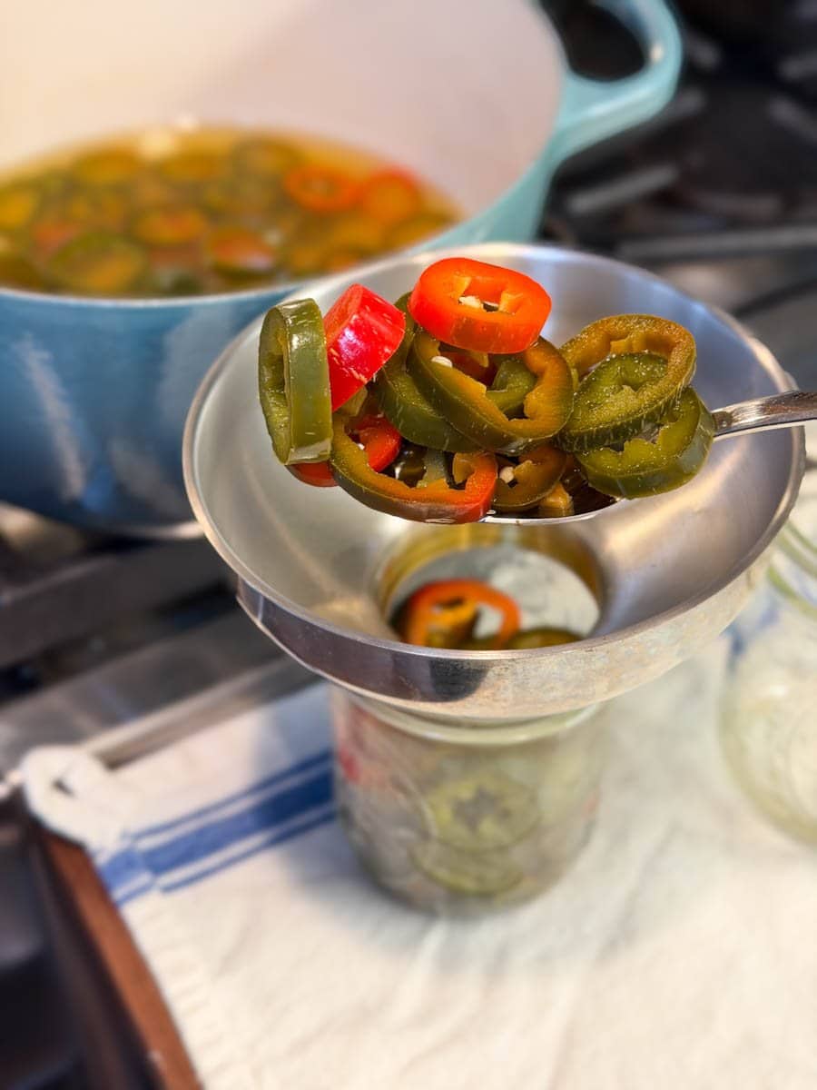A large slotted spoon is being used to add pickled jalapeños to pint jars with a metal canning funnel.