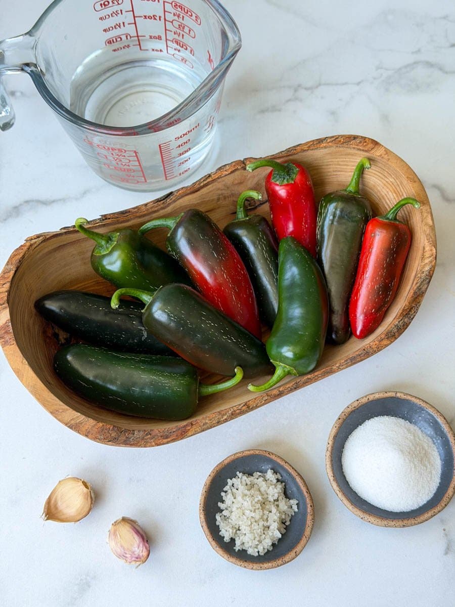 An oblong oval wood bowl is full of green chili peppers. A few pinch bowls are each full of salt and sugar while two cloves of garlic and a liquid measuring cup of vinegar and water sit nearby.