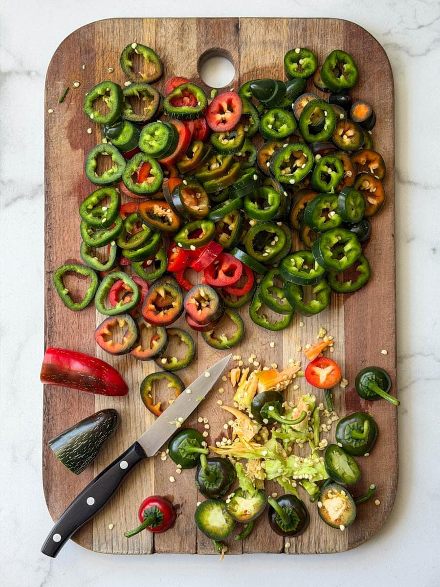 Sliced jalapeños are being processed on a wood cutting board. A small pairing knife is next to the tops and cores of the peppers on one side of the cutting board.