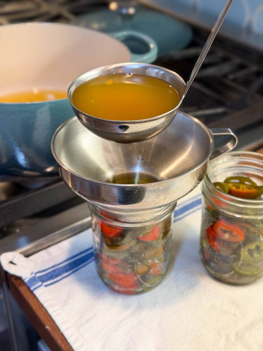 A ladle is being used to spoon brine into pint jars of pickled jalapeños using a metal canning funnel.